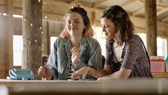 在餐厅的野餐桌上，两个女孩分享着耳机，随着音乐和鼓声起舞。视频素材