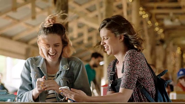 两个女孩一起看智能手机，在有风的野餐棚里笑。视频素材