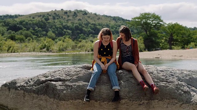 阳光明媚的日子，两姐妹坐在俯瞰河流的岩石上拥抱。视频素材