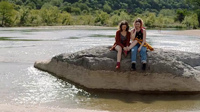 在阳光明媚的日子里，两个女孩在徒步旅行中穿过岩石，互相倚靠着坐着俯瞰河流。视频素材
