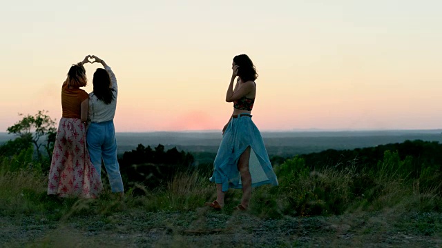 在神奇的时刻，在风景优美的山腰上，穿着永恒连衣裙的女孩们双手做成心形。视频素材