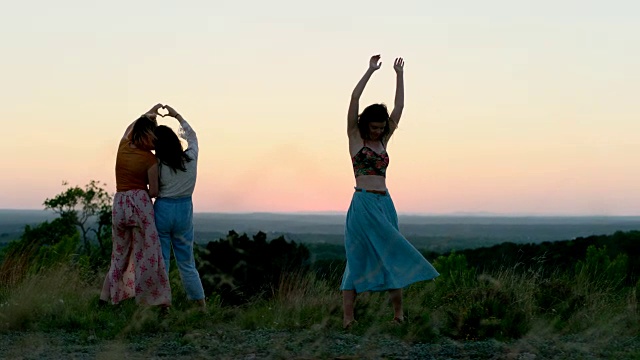 日落时分，女孩在山顶上跳舞，朋友们用手做成心形。视频素材