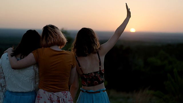 女孩和朋友们站在山坡上，手在空中挥手，看着夕阳下的绿色山谷。视频素材
