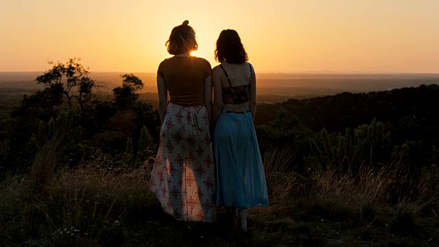 两个穿着飘逸连衣裙的女孩从绿色的山腰观看山谷中的日落。视频素材