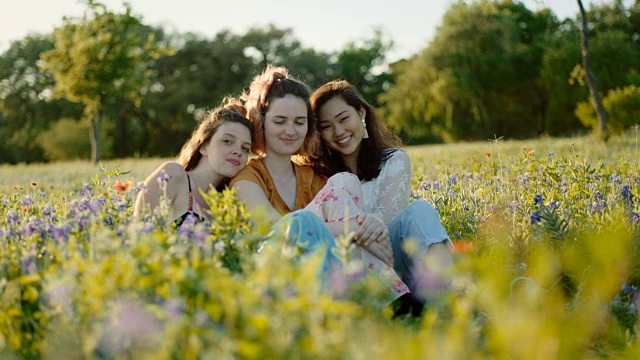 三个女孩坐在花田里，靠着对方，对着镜头微笑。视频素材