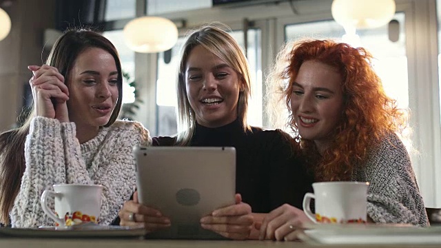 三名年轻女子在咖啡馆里一边笑一边用平板电脑交流。视频素材