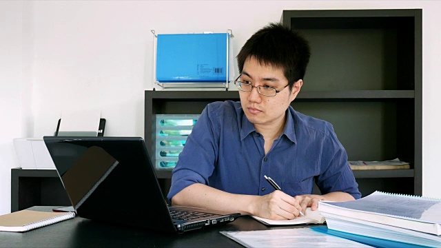 戴眼镜的亚洲商人在家庭办公室用笔记本电脑和手机工作视频下载