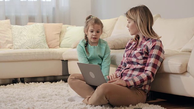 年轻的母亲和她的小女儿一起在地毯上休息时使用笔记本电脑。视频素材
