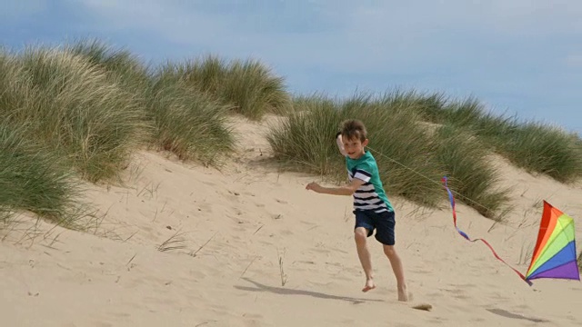 一个小男孩有很大的乐趣沿着海滩沙丘跑放他的风筝。视频素材