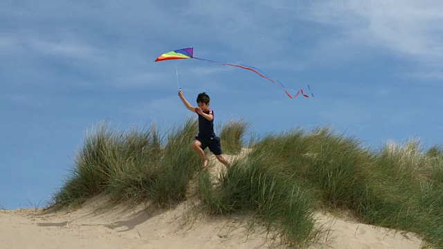 一个小男孩在放风筝，他从一个沙丘上跳下来，笑着玩。视频素材