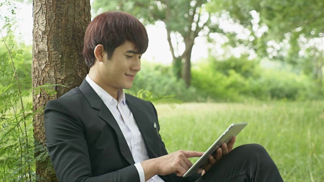 亚洲年轻男性西装在公园使用平板电脑视频下载