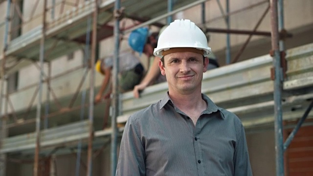 一位戴着白色头盔的男建筑师在施工现场滚动着平面图，微笑着对着摄像机视频下载