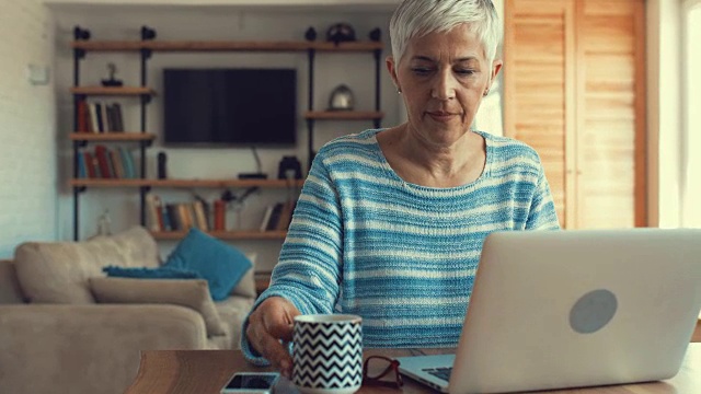 成熟的女人在家里一边喝咖啡一边用笔记本电脑。视频素材