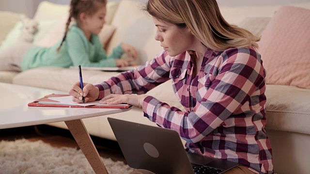 年轻的母亲在家里工作时使用笔记本电脑并做笔记。视频素材