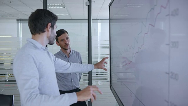 两个白人男同事在讨论会议室大屏幕上显示的一个财务图表视频素材