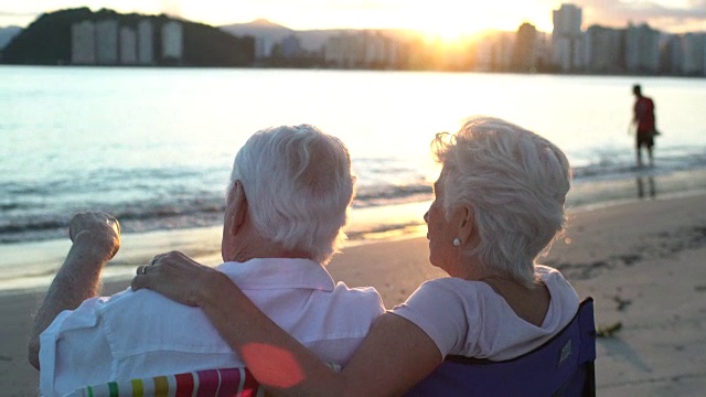 移动手持相机从一个老夫妇坐在沙滩椅子上的背影观看日落与一个集市的镜头。视频下载