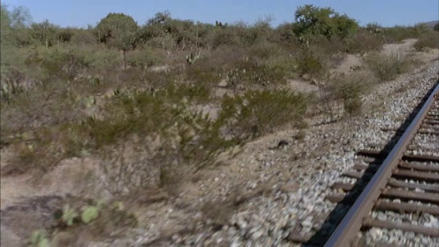 中等角度的铁轨在镜头下快速通过。看火车轨道上的水平木板。看到路边的砾石和绿色的灌木丛。可能在沙漠地区。视频下载