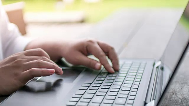 女人用手在笔记本电脑键盘上打字，以绿色的散焦为背景，时间流逝运动，缩小运动视频下载