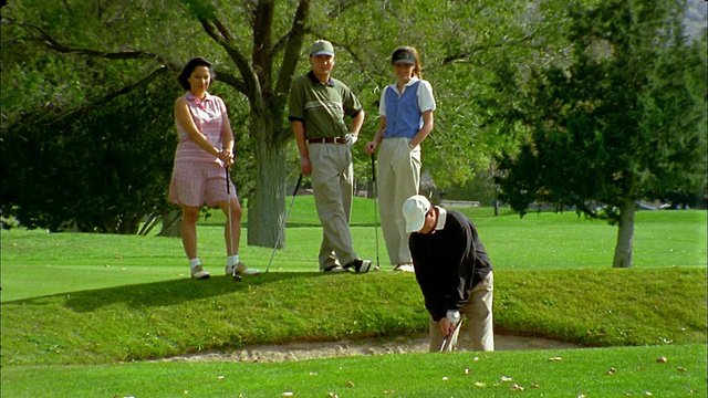 一名中枪男子在沙坑里打高尔夫球，背景中有一名女子和另一对夫妇在观看视频素材