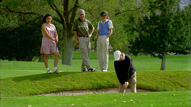 一名中枪男子在沙坑里打高尔夫球，背景中有一名女子和另一对夫妇在观看视频素材