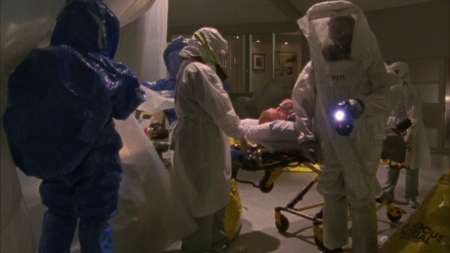 中等角度的急救室团队或工作人员穿着危险生物防护服。戴着防毒面具的男男女女从头到脚都穿着白色和浅蓝的套装。被推到担架、轮床或病床上的人。大的黄色有害物质视频下载