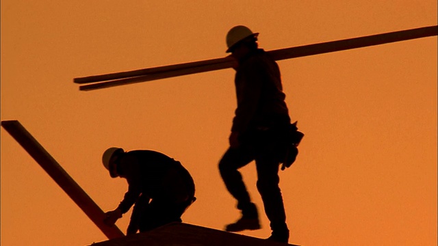 两个建筑工人在屋顶上搬运木板的中景剪影/凤凰城，亚利桑那州视频素材