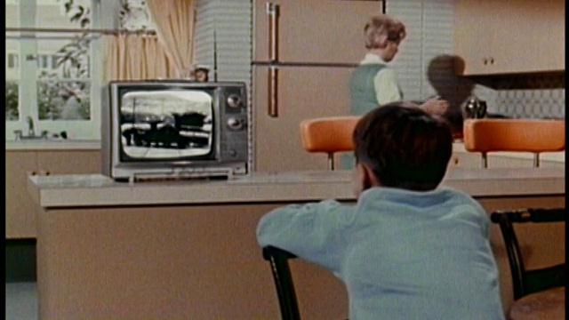 20世纪60年代，一个中等镜头的男孩在厨房柜台上看电视，他的母亲在背景/音频中视频下载