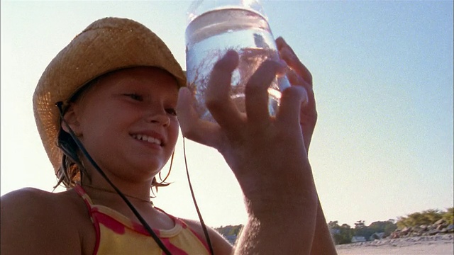 低角度近距离拍摄年轻的女孩在草帽和微笑着拿着罐子/植物在海滩上视频素材