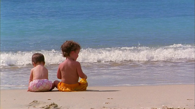 中镜头后视图两个小孩坐在海滩上，看着海浪/孩子转向CAM视频素材