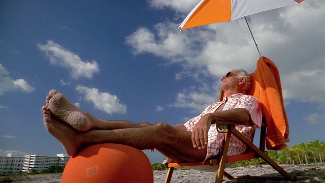 低角度中拍摄的高级男子坐在伞下沙滩椅，他的脚在脚休息，以蓝天为背景视频下载