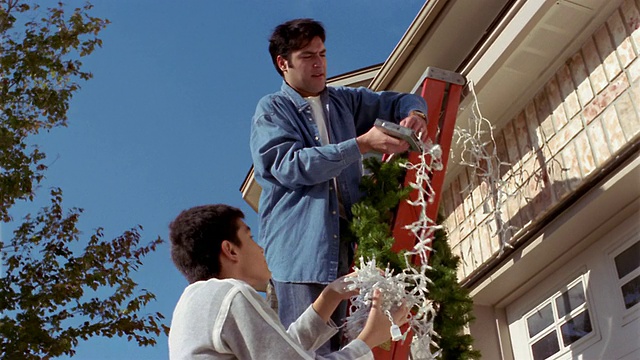 低角度中拍摄父亲和儿子在梯子上，挂在房子上的圣诞装饰品视频下载