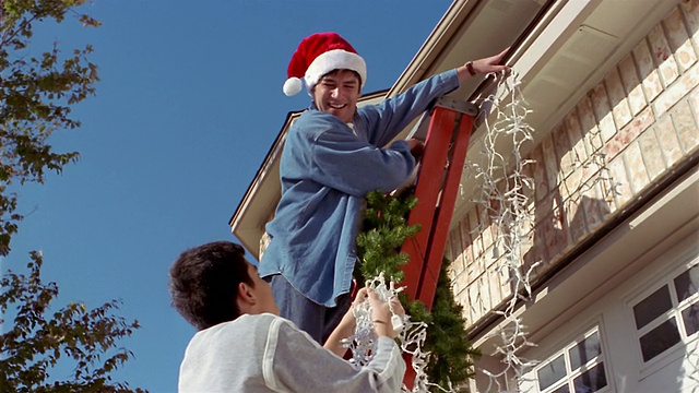 低角度中拍摄父亲(戴着圣诞帽)和儿子在梯子上，挂在房子上的圣诞装饰品视频下载