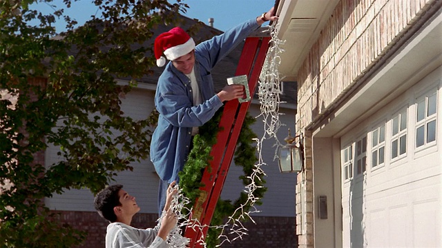 中拍父亲(戴着圣诞帽)和儿子在梯子上，把圣诞装饰品挂在房子上视频下载