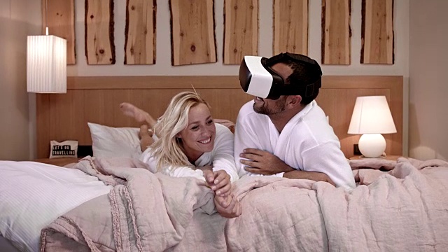这对30多岁的情侣一起躺在酒店房间的大床上，穿着白色浴袍，他留着黑色短发和修剪过的胡子，一边玩一边戴着VR眼镜进行360°虚拟现实游戏体验，金发女孩最后厌倦了视频下载