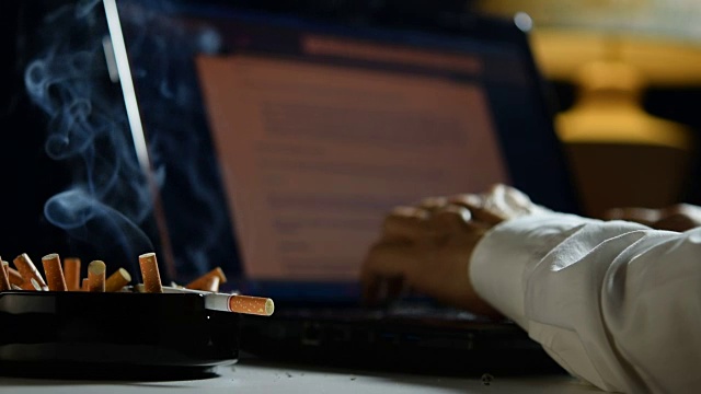 一个男人在笔记本电脑上工作，在一个装满烟头的烟灰缸上抽烟视频下载