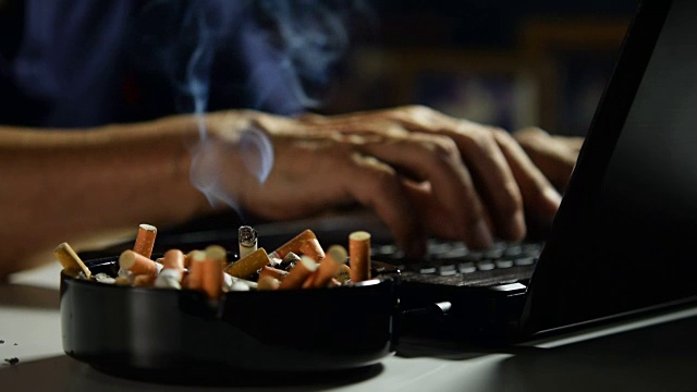 画面中，一名男子用手指夹着点燃的香烟，在用笔记本电脑工作视频下载