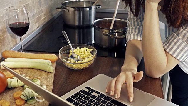 年轻女子一边在厨房做饭，一边在笔记本电脑上读菜谱。视频下载