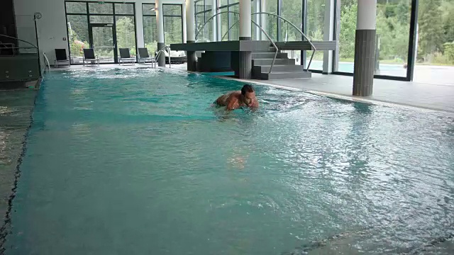 在酒店SPA的室内泳池区域，白天有周围的全景窗——一个30多岁的男人，留着黑色短发，穿着绿松石泳衣，潜入蓝色的水中，以蛙泳的姿势游向相机，走出水，走出画面视频下载