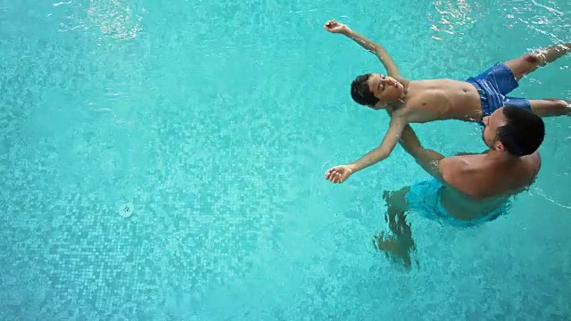室内游泳池-晒得黝黑的男人在他的30多岁短发和他的8岁前的儿子一起在绿松石蓝色的热水，单身父亲做水平衡他的孩子，孩子是漂浮在水上面的镜头视频下载