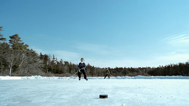长镜头男孩滑冰冰球在冰冻的池塘前景/打冰球的朋友在背景/缅因州视频下载