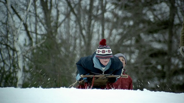 中镜头男孩骑着雪橇从白雪覆盖的山上滑下来/父母在后面看着视频下载