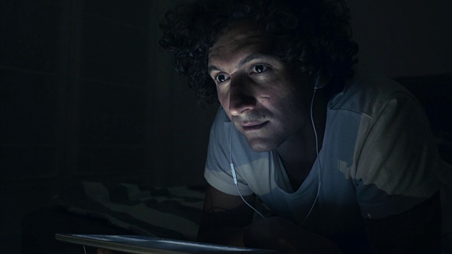 年轻人在床上戴着耳机用数码平板电脑。视频素材