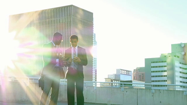两个微笑的商人站在停车场的屋顶上看智能手机上的数据视频素材