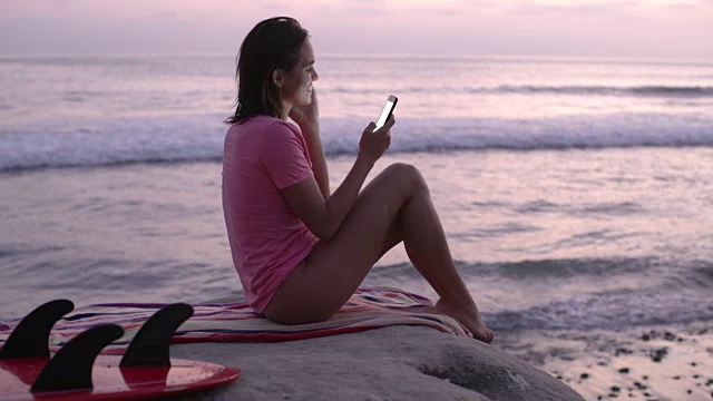 年轻女子坐在海边用她的手机视频下载