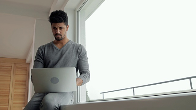 年轻的黑人坐在家里的窗户边，使用笔记本电脑。视频素材