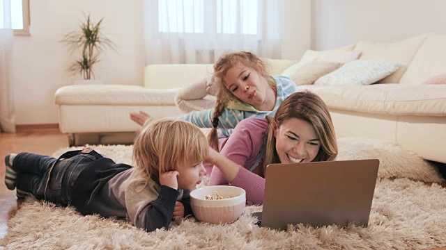 快乐的妈妈和她的两个孩子使用笔记本电脑，在客厅吃爆米花。视频素材