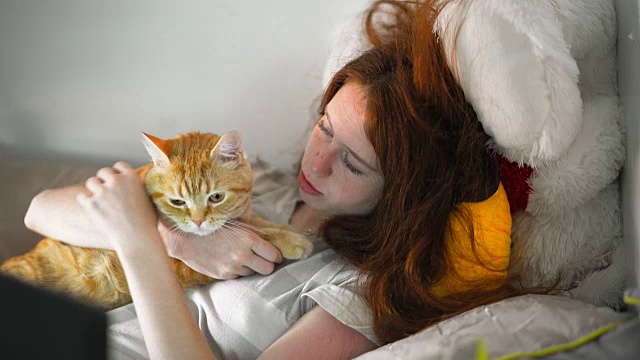 一个十几岁的女孩在抚摸着躺在床上的红色虎斑猫视频素材