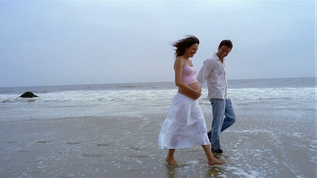 广角镜头孕妇和男人牵着手+走过海滩冲浪/女人揉肚子视频素材