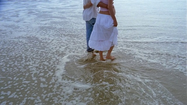 中镜头倾斜的孕妇和男人赤脚通过冲浪在海滩上手臂环抱视频素材
