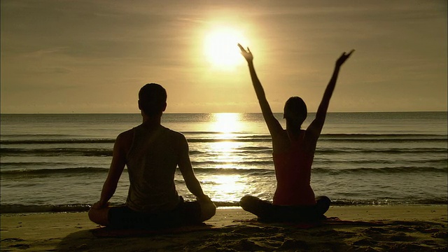 男人和女人在冥想，坐在瑜伽莲花姿势，和女人伸展在日出的海滩上/华欣，泰国视频下载